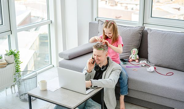 Mann sitzt entspannt im Wohnzimmer am Computer. Tochter spielt bei ihm.